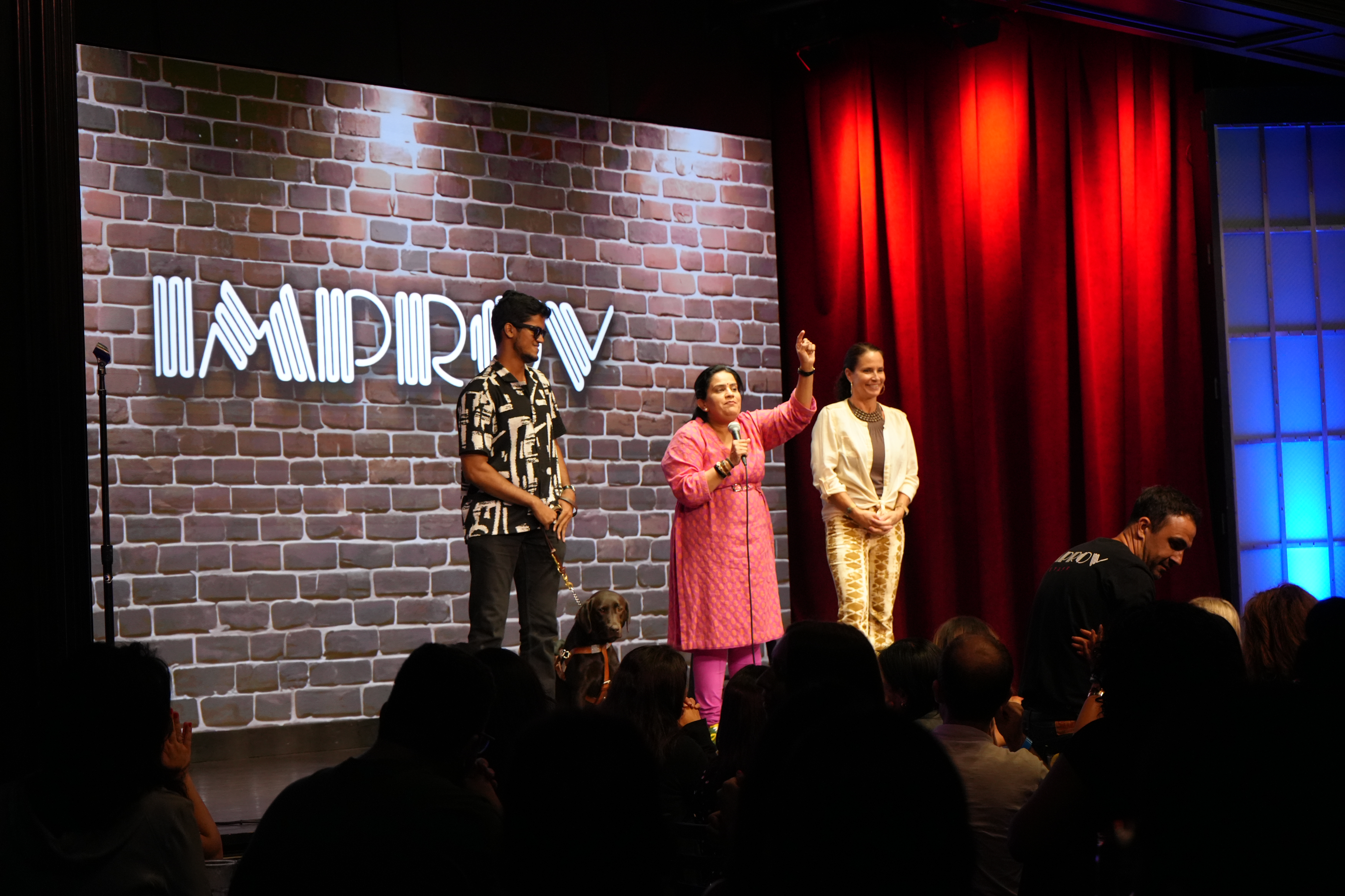 Left to right: Bhavya Shah, Zarna Garg, and Mary Capone stand beneath the glowing Improv logo as Zarna addresses an applauding audience